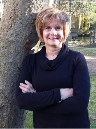 A picture of Dr. Mason leaning against a tree. She has short, blonde hair and is wearing a black, long-sleeved shirt.
