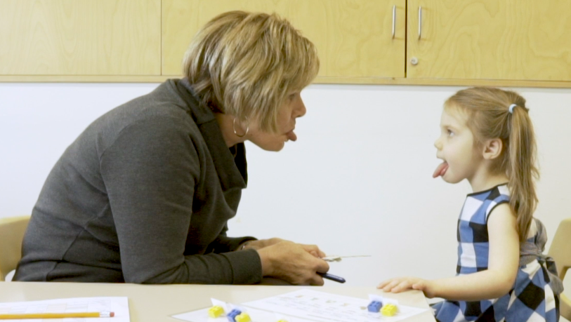 Dr. Mason and a little girl face each other and stick their tongues out, demonstrating a task in the study.