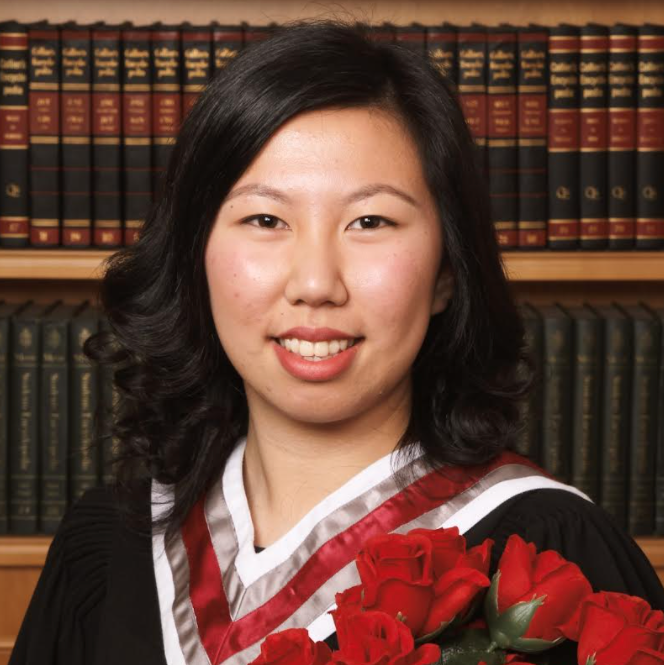 Hannah has medium, curled black hair, is wearing a graduation gown, is holding a bouquet of red roses, and is smiling.